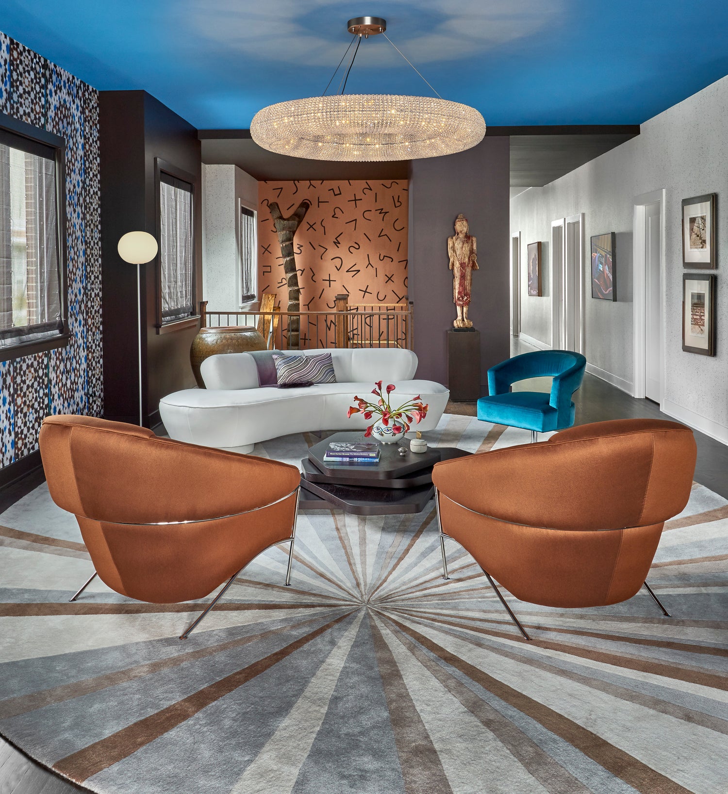A luxury modern living room featuring two prominent rust-brown velvet armchairs in the foreground, a white curved sofa, a bright blue accent chair, and a large angela adams Jimi geometric patterned area rug. The room has a bold blue ceiling, a crystal chandelier, and various decorative sculptures.