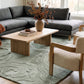A modern living room setup featuring a dark gray sectional sofa, a light wood coffee table, two cream-colored armchairs with wood frames, and the Nebula Dream SuperLuxe sage green area rug on a light wood floor.
