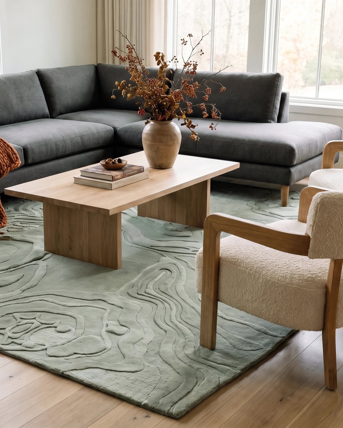 A modern living room setup featuring a dark gray sectional sofa, a light wood coffee table, two cream-colored armchairs with wood frames, and the Nebula Dream SuperLuxe sage green area rug on a light wood floor.