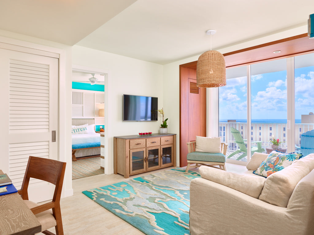 A bright, living room at the margaritaville residences in Florida, featuring the angela adams ocean seaglass turquoise area rug, wooden cabinet, and a large window overlooking the ocean. A wicker pendant light hangs above, and a doorway in the background reveals a bedroom with turquoise bedding.