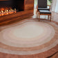 The angela adams Pool Heaven organic-shaped area rug with concentric oval bands in shades of beige, cream, tan, and medium brown, styled on a dark wood floor in a modern living room with a fireplace and a black leather chair.