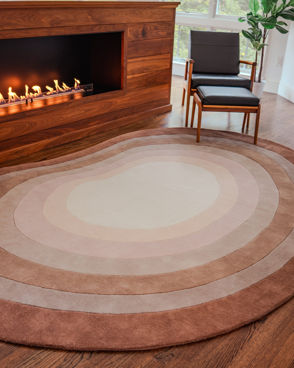 The angela adams Pool Heaven organic-shaped area rug with concentric oval bands in shades of beige, cream, tan, and medium brown, styled on a dark wood floor in a modern living room with a fireplace and a black leather chair.