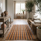 Rustic entryway with a plaid rug, wooden console table, potted olive tree, and bench in a warm, minimalist home interior.