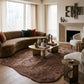 Modern living room with a curved brown velvet sofa, textured floral rug, brass coffee table, and minimalist fireplace.