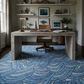 Coastal home office with a wave-pattern area rug, light wood desk, rattan chair, and nautical wall art on shiplap walls.