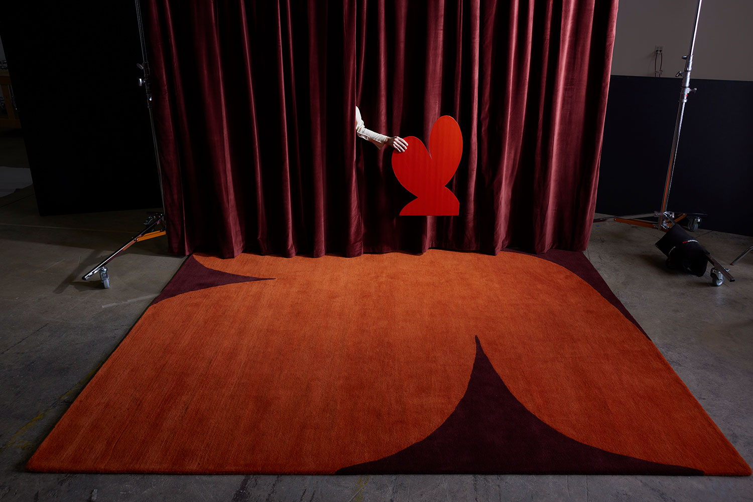 A hand behind a stage curtain places a cut out heart above a modern, hand tufted area rug called, Dove Love by Angela Adams
