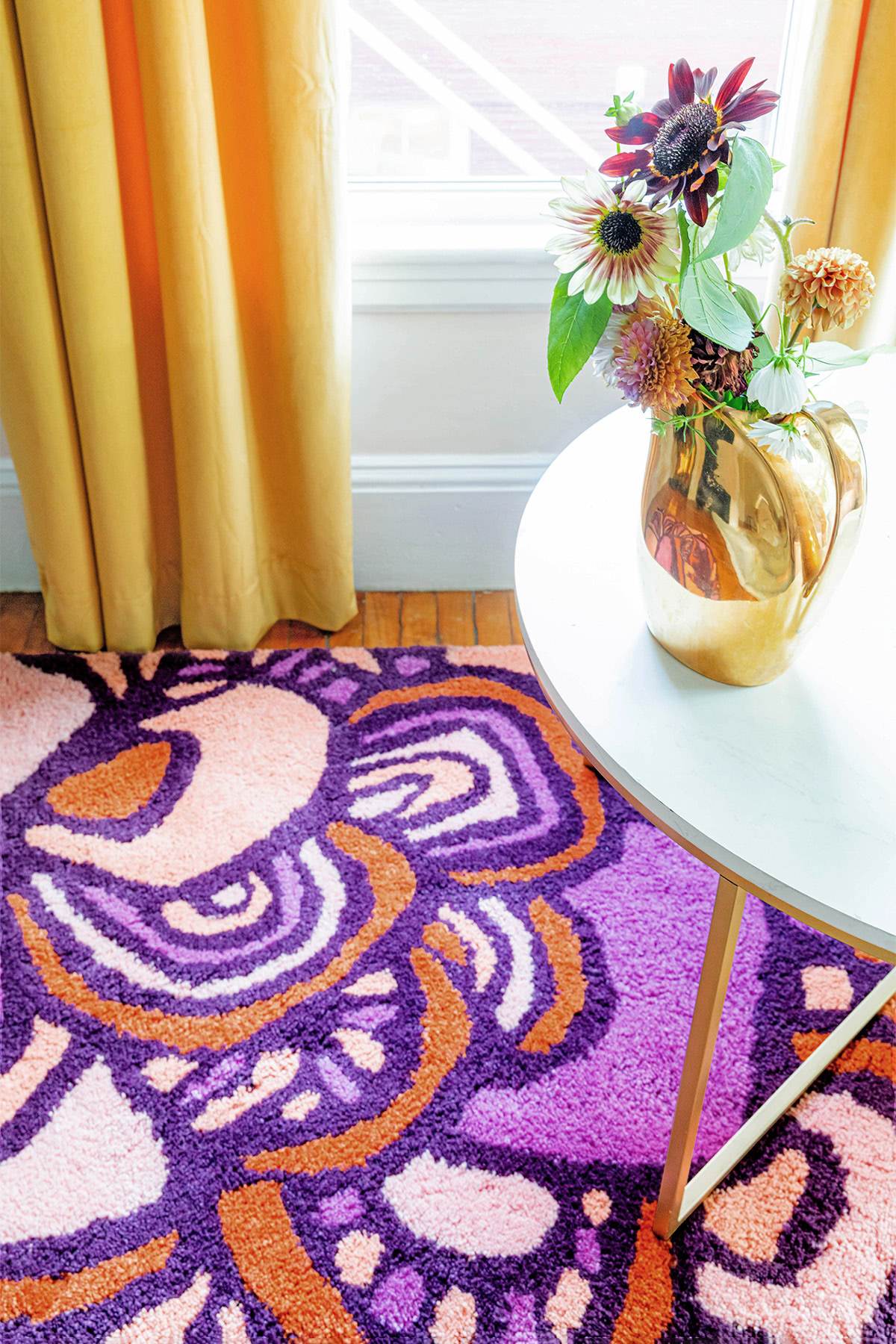 A sunny room with flowers on a table sitting on a red, and pink floral pattern, modern area rug called Maeve Tulip