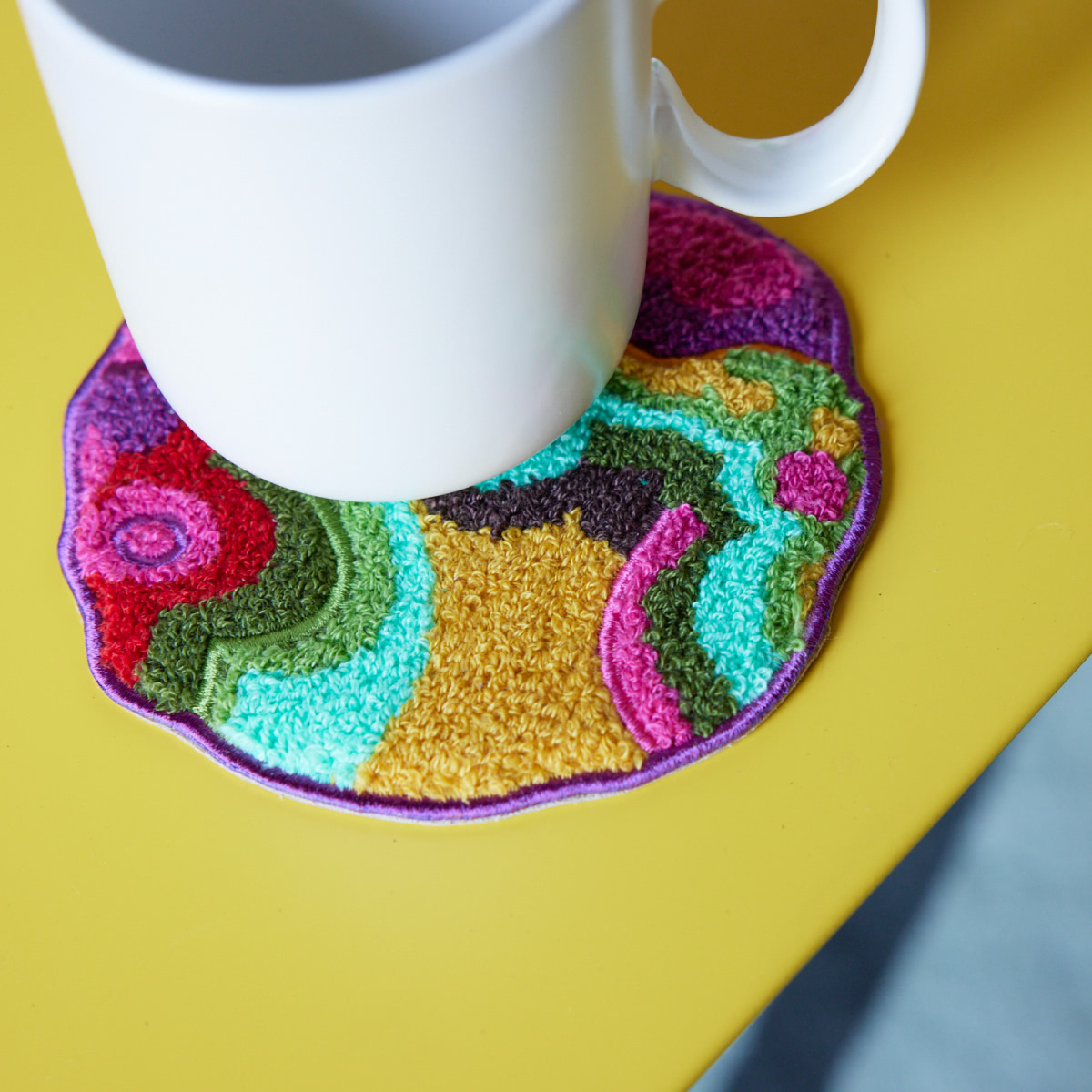 A white mug rests on a miniature rug called Mini Oasis, in Garden color way by Angela Adams.