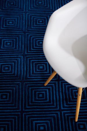 A wooden chair sits on a modern area rug called Duke Blue by Angela Adams